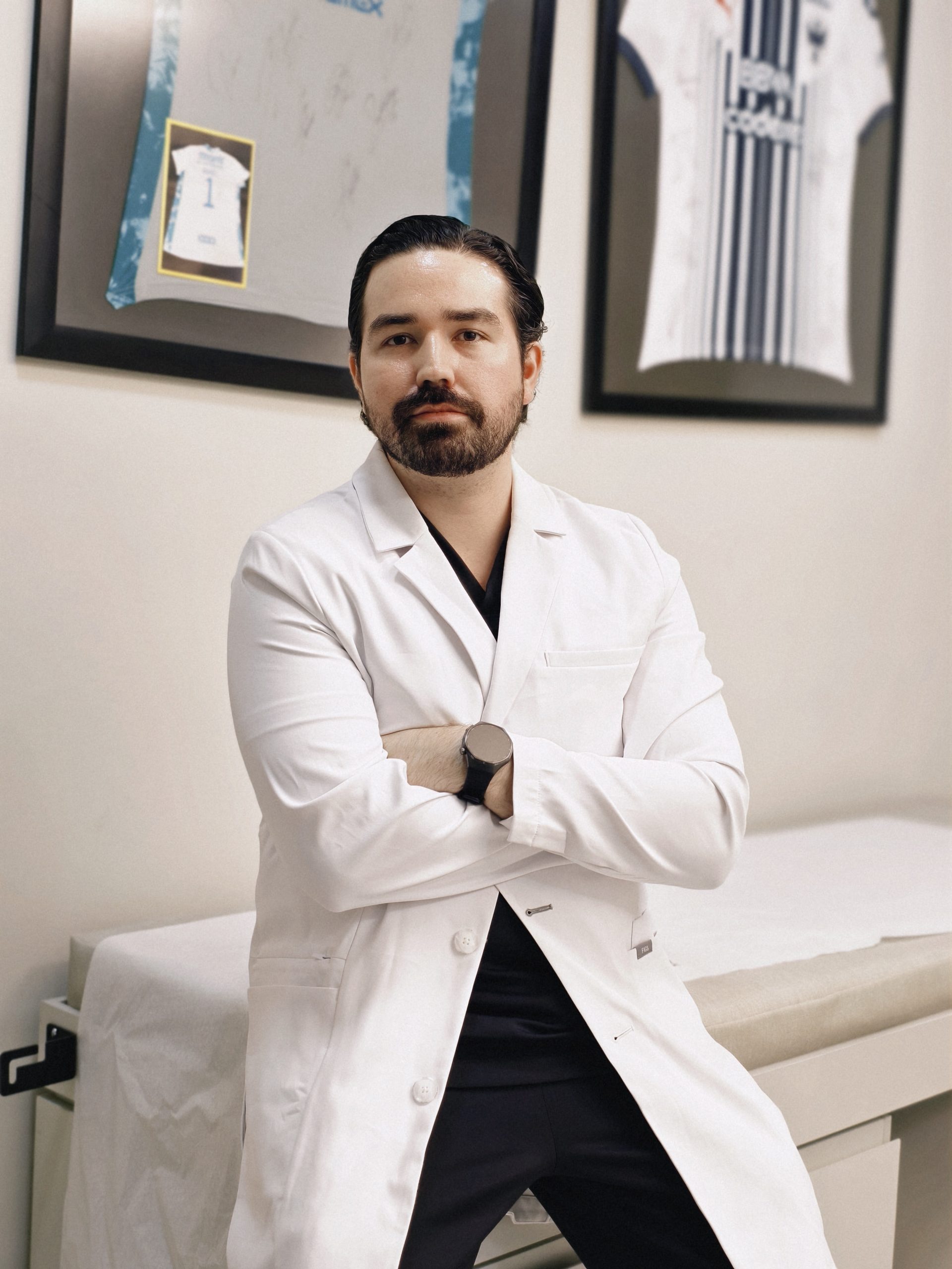Dr. Braulio Ayala en su consultorio de Doctors Hospital, Monterrey, con jerséis deportivos enmarcados de fondo
