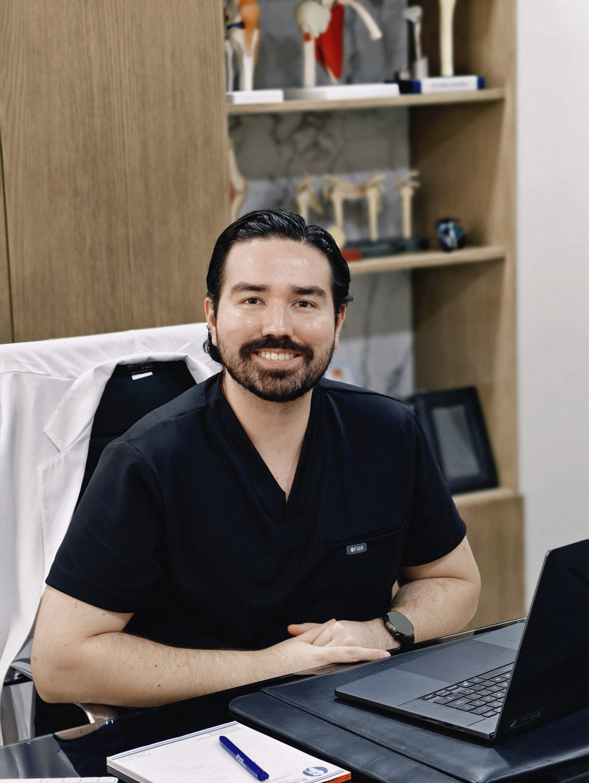Dr. Braulio Ayala sonriendo en su consultorio, junto a modelos anatómicos y su laptop